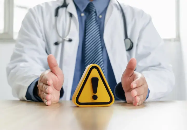 Medico con camice bianco e stetoscopio, mani aperte su un segnale di pericolo. Attenzione alle raccomandazioni ministeriali per la salute.