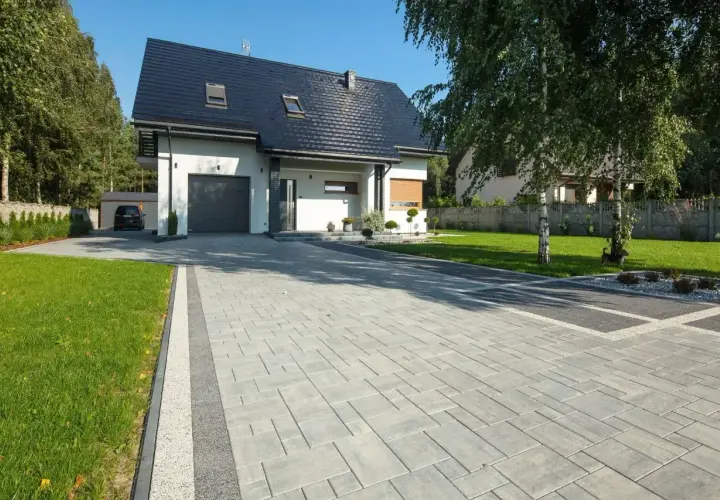 Nowoczesny podjazd przed domem z szarej kostki brukowej, otoczony zielonym trawnikiem i drzewami.