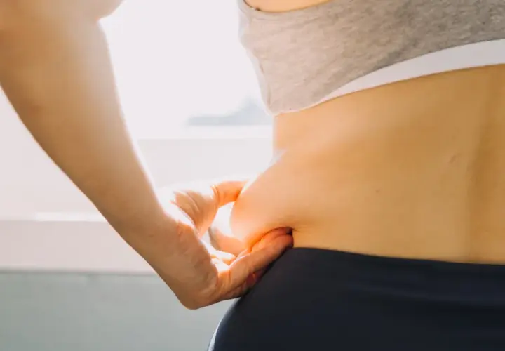 Dłoń chwyta fałd skóry na boku, symbolizując troskę o sylwetkę i chęć poznania, jak przyspieszyć metabolizm.