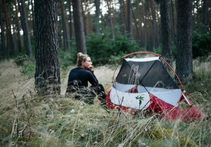 Ein Mann sitzt nachdenklich neben einem roten Zelt im Wald. Hier darf man im Wald zelten und die Natur genießen.