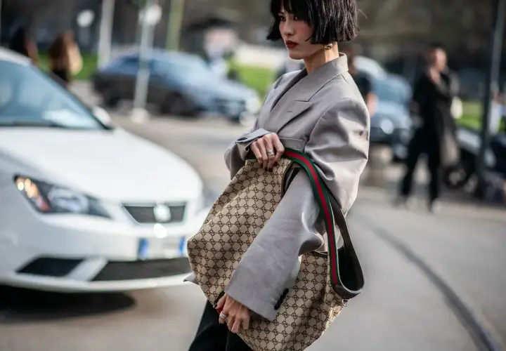 Kobieta w szarym żakiecie trzyma torbę z monogramem Gucci, jakby to był certyfikat autentyczności.