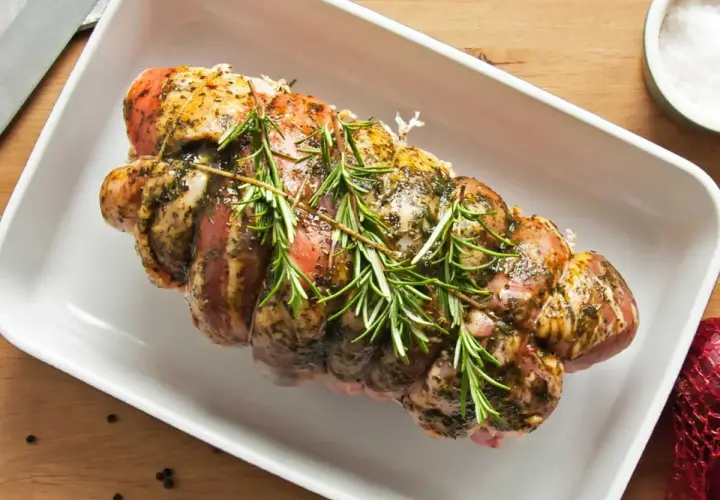 Un rôti de porc rosé, assaisonné et garni de romarin frais, prêt à être cuit dans un plat blanc.