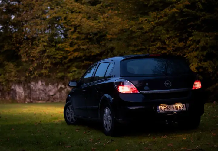 Czarny Opel Astra H 1.6 benzyna zaparkowany na trawie, w tle jesienne drzewa.