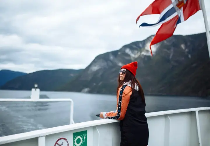 Kobieta na statku pod norweską flagą podziwia fiordy. Podróż po Norwegii, mimo że piękna, może wiązać się z wysokimi cenami.