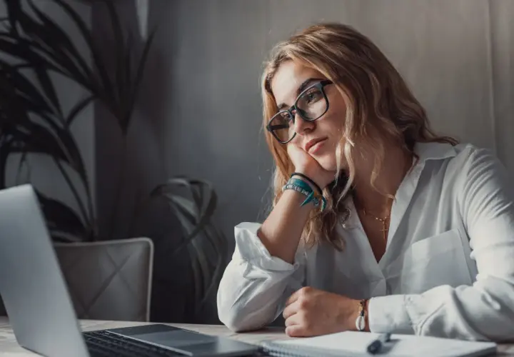 Kobieta w okularach opiera głowę na dłoni, patrząc zamyślona na laptopa.