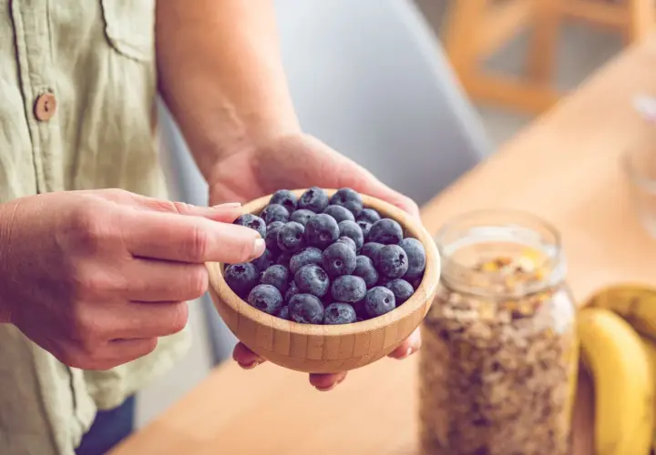 Hände halten eine Schale voller Blaubeeren, reich an Flavonoiden. Daneben stehen Müsli und Bananen – gesunde Flavonoide Lebensmittel für den Start in den Tag.