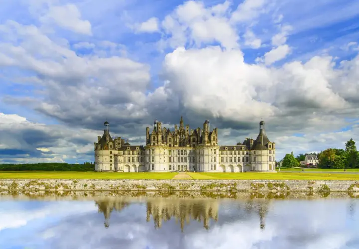 Zamek Chambord nad Loarą odbijający się w wodzie, pod burzliwym, błękitnym niebem.