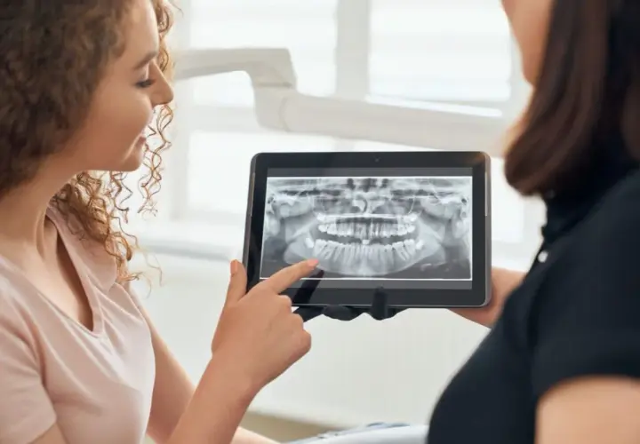 Pacjentka z kręconymi włosami ogląda z dentystką panoramiczny rentgen stomatologiczny na tablecie.