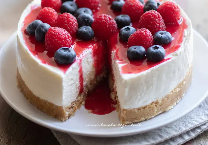 Cheesecake alla frutta con lamponi e mirtilli freschi, ricoperta da una glassa rossa lucida. Una fetta è stata tolta, rivelando la morbida crema.