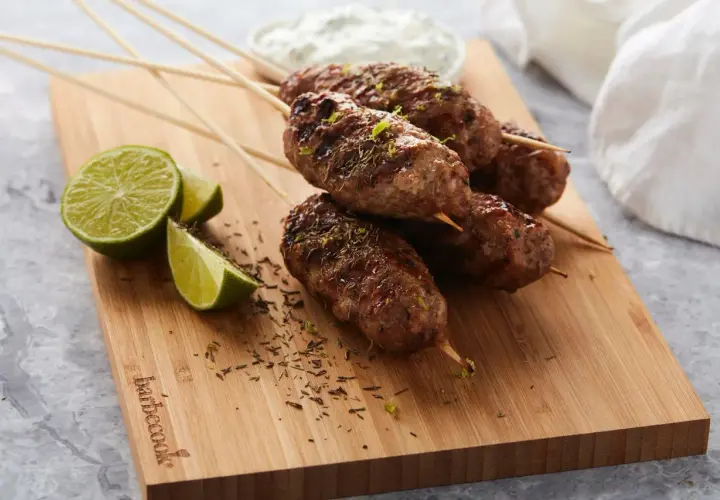 Brochettes d'agneau grillées, garnies d'herbes et de zeste de citron vert, servies avec des quartiers de citron vert et une sauce blanche.