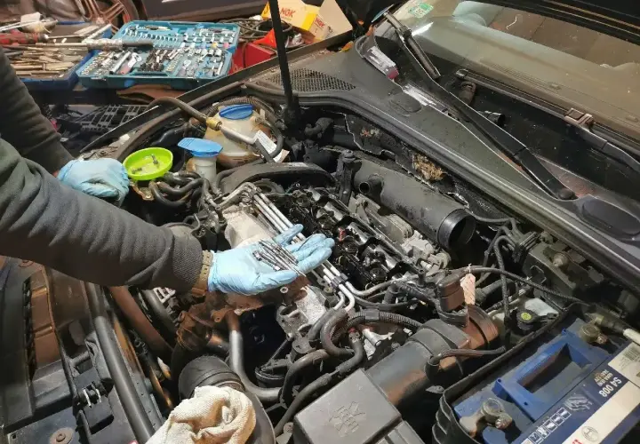 Mechanik w niebieskich rękawiczkach trzyma świece żarowe do wymiany. W tle skrzynka z narzędziami i silnik samochodu.