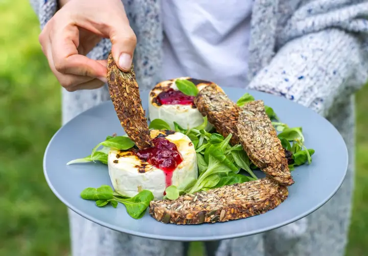 Ręka zanurza chrupiący sucharek w grillowanym serze z konfiturą. Czy takie sucharki są kaloryczne? Zależy od składu, ale te pełne ziaren wyglądają zdrowo!