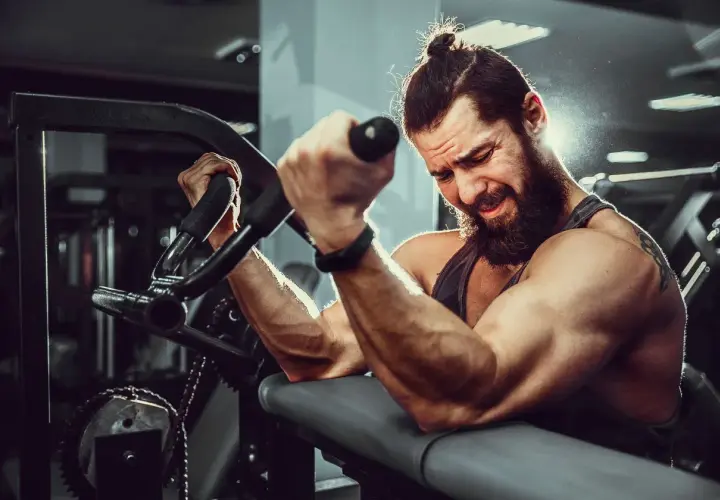 Mężczyzna z brodą i kitką ćwiczy biceps na maszynie. Widać jego napięte mięśnie i determinację. To idealne miejsce, by budować **mutant mass sklad**.