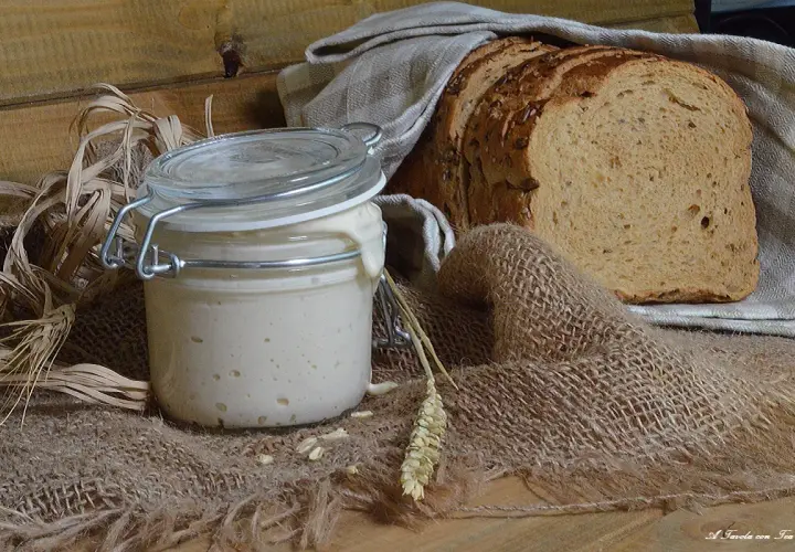 Un barattolo di lievito madre attivo, con una fetta di pane rustico sullo sfondo. Ingredienti naturali per un pane fragrante.