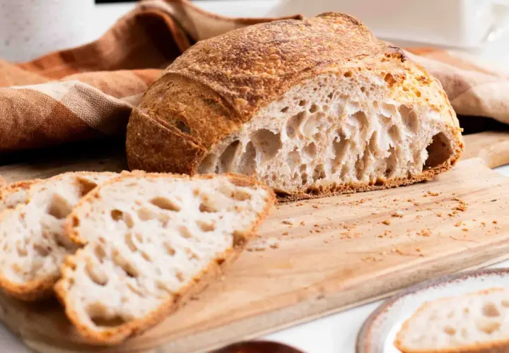 Un delicioso pan de masa madre, con su corteza dorada y miga aireada, reposa sobre una tabla de madera junto a unas rebanadas.