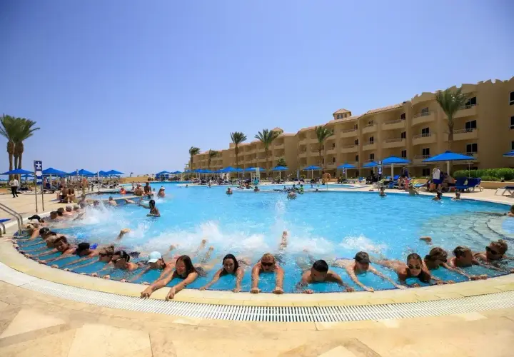 Grupa ludzi pływa w basenie hotelu, tworząc falę. W tle budynek hotelu i palmy. Wiele pozytywnych amwaj blue beach resort & spa opinie potwierdza ten widok.