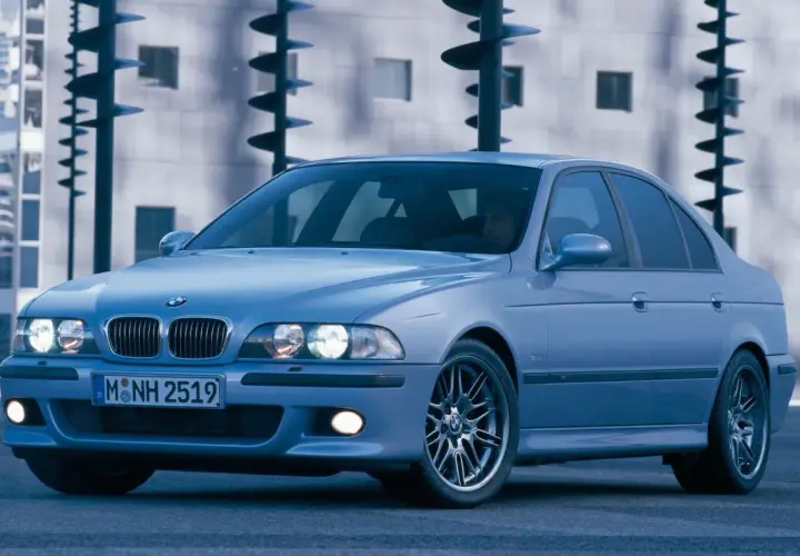 Błękitne e39 bmw z felgami M, zaparkowane przed nowoczesnym budynkiem.
