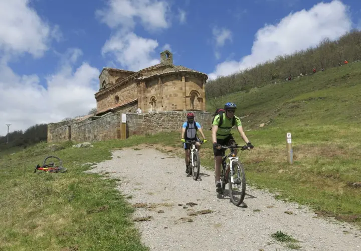 Ciclistas exploran el románico palentino, pedaleando hacia una iglesia de piedra bajo un cielo azul con nubes.