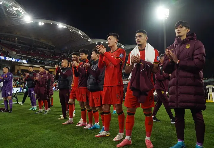 Piłkarze w czerwonych strojach, być może reprezentacja Chin, klaszczą na boisku. W tle widać trybuny i światła stadionu.