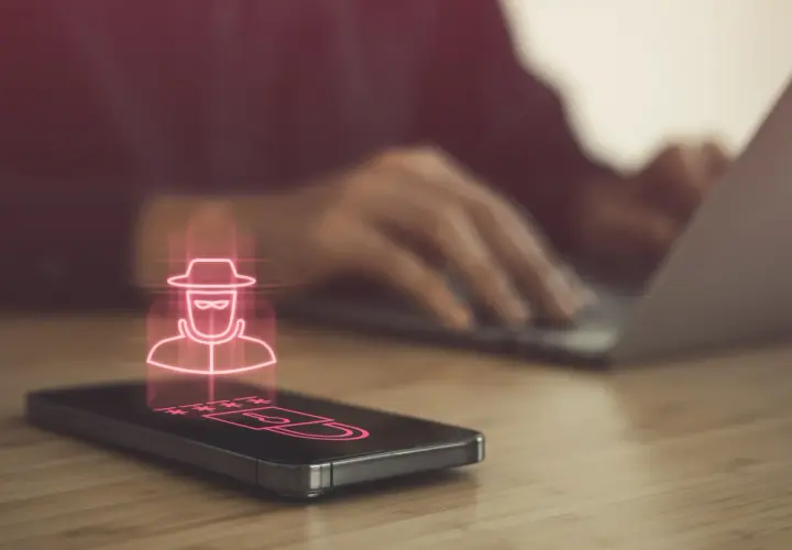 Haker w kapeluszu i gwiezdki symbolizujące hasło na ekranie telefonu, sugerujące podsłuch w telefonie. W tle osoba pracuje na laptopie.
