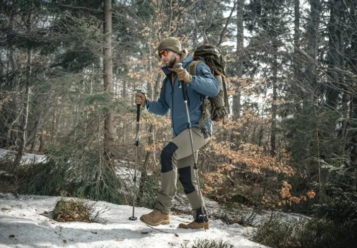 Mężczyzna w zimowej kurtce i czapce idzie przez las, wspierając się na kijach trekkingowych. W tle drzewa i śnieg.