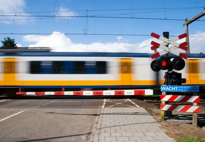 Pędzący pociąg mija przejazd kolejowy z czerwonym światłem i znakiem "Wacht". Widoczny jest charakterystyczny krzyż Andrzeja.