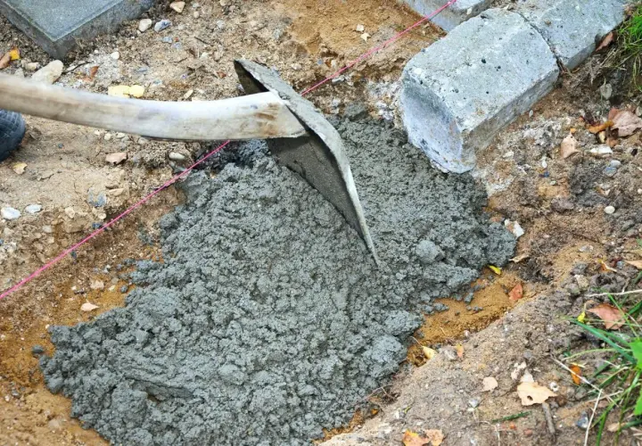 En hacka sprider jordfuktig betong i en grop. Betongblock ligger bredvid.