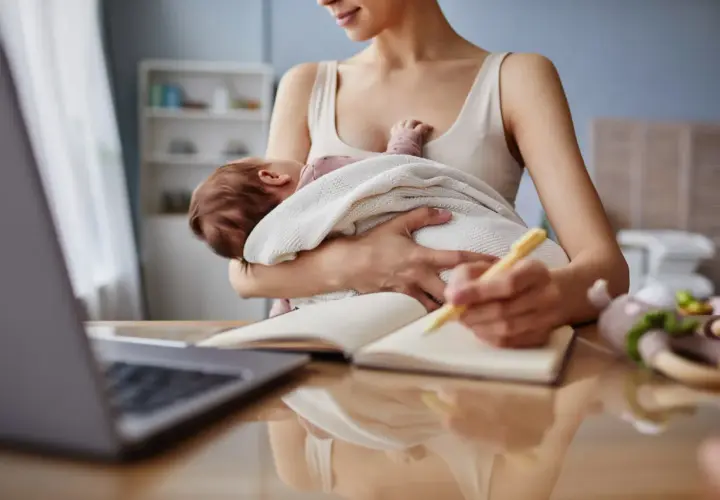 Kobieta trzyma dziecko i pisze w notatniku przy laptopie.