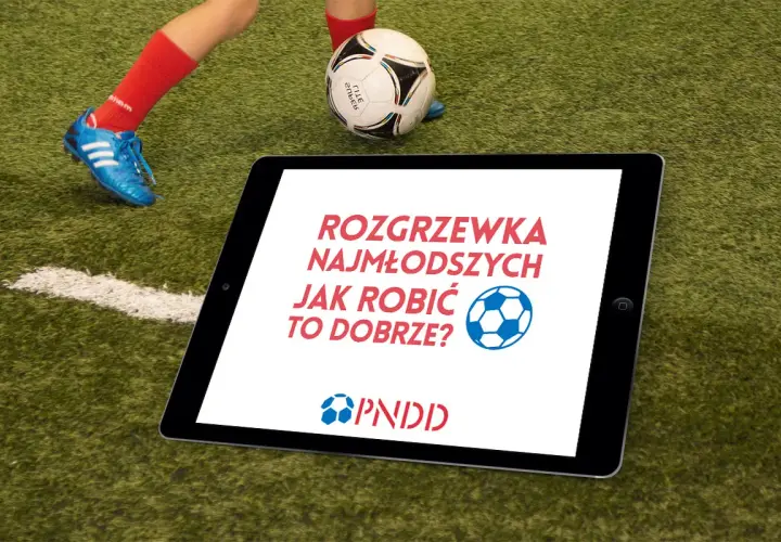 Tablet na boisku z napisem "Rozgrzewka najmłodszych jak robić to dobrze?". Obok piłka i noga dziecka w czerwonej skarpecie.