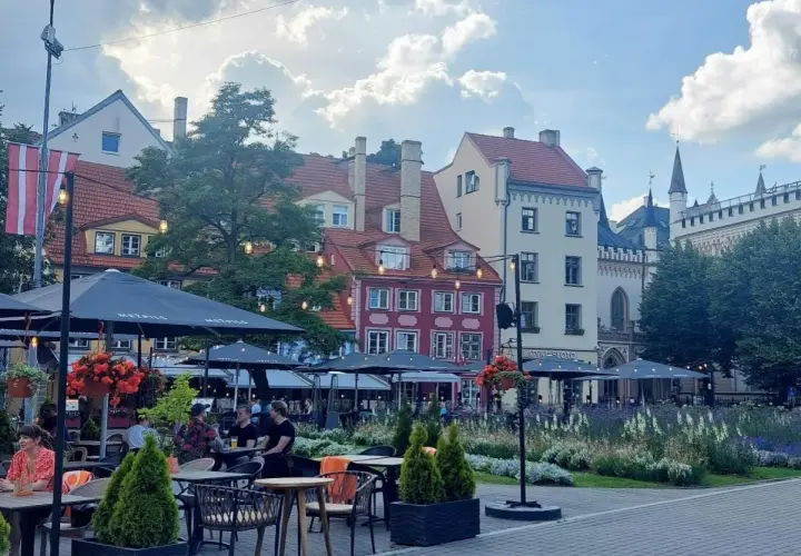 Tętniący życiem plac w Rydze, z kolorowymi kamienicami i kawiarnianymi ogródkami. W tle widać fragmenty zabytkowej architektury, przypominającej o historii państwa z Kurlandią.