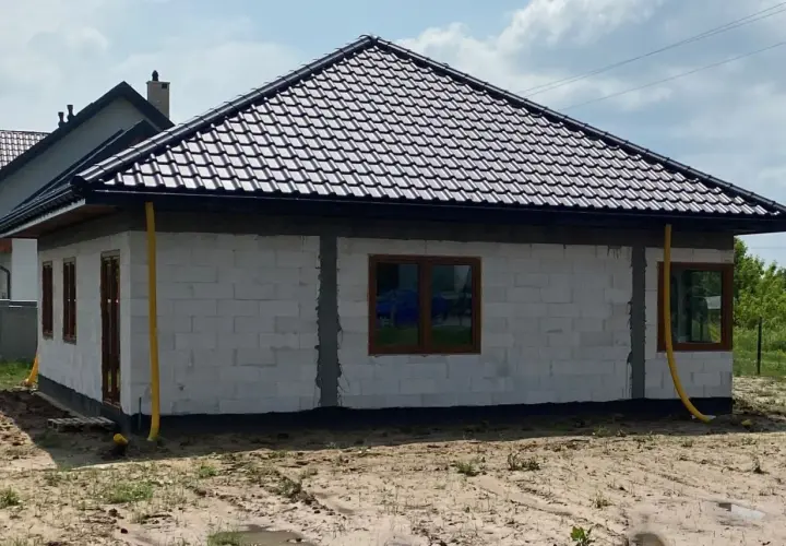 Dom w budowie z żółtymi rurami odprowadzającymi wodę z rynien. Widać białe bloczki, brązowe okna i dach pokryty ciemną dachówką.
