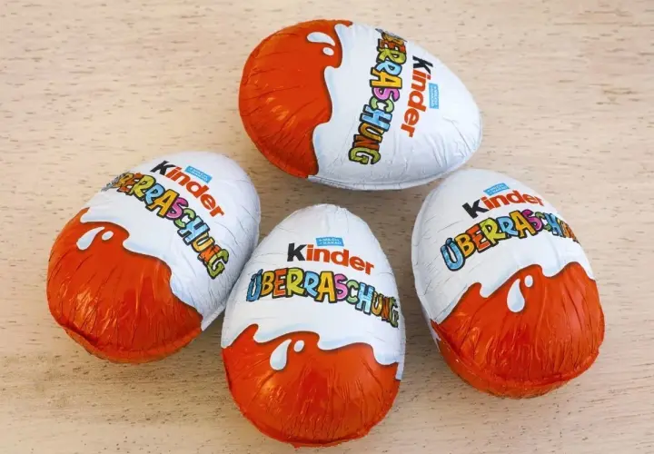 Cztery jajka Kinder Niespodzianka w pomarańczowo-białych opakowaniach na drewnianym tle.