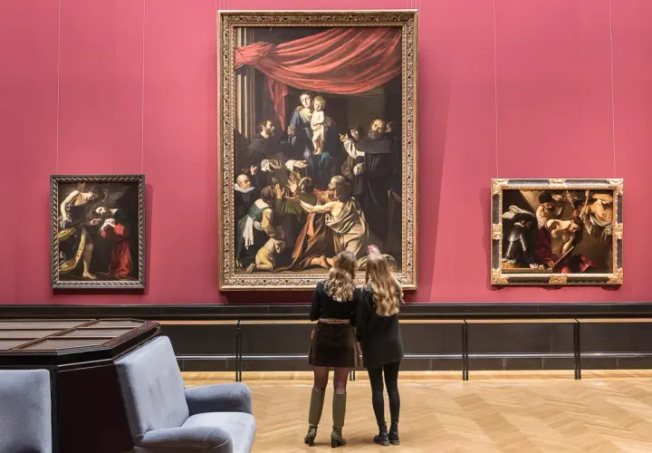Dwie kobiety oglądają obrazy w muzeum sztuki na tle czerwonej ściany.
