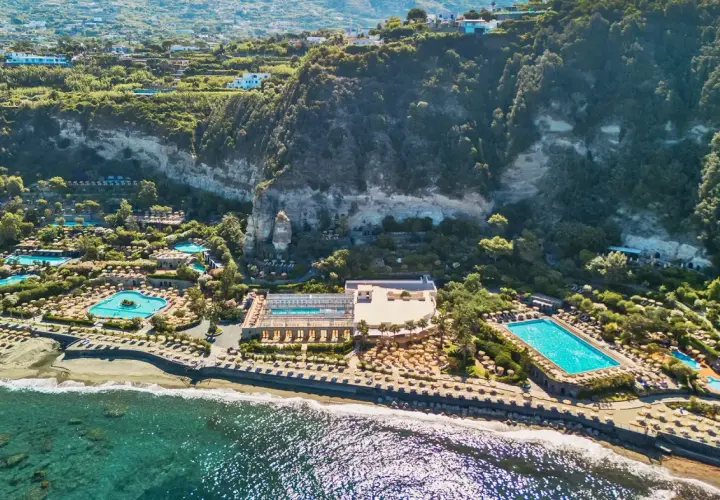 Vista aerea di lussuosi complessi termali a Ischia, con piscine scintillanti, lettini e vegetazione rigogliosa che si affacciano sul mare.