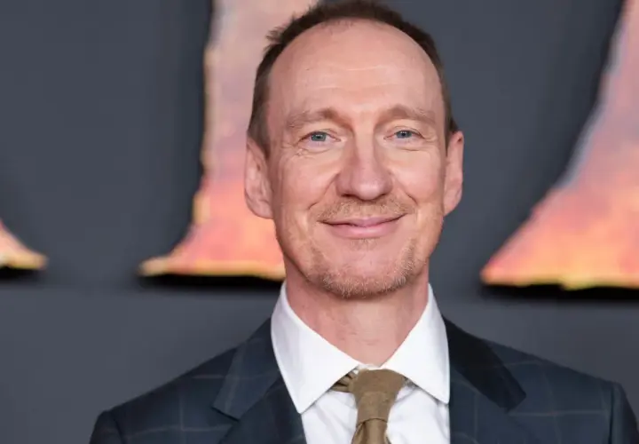 Uśmiechnięty David Thewlis w garniturze i krawacie, z niebieskimi oczami, na tle rozmytych, pomarańczowych kształtów.