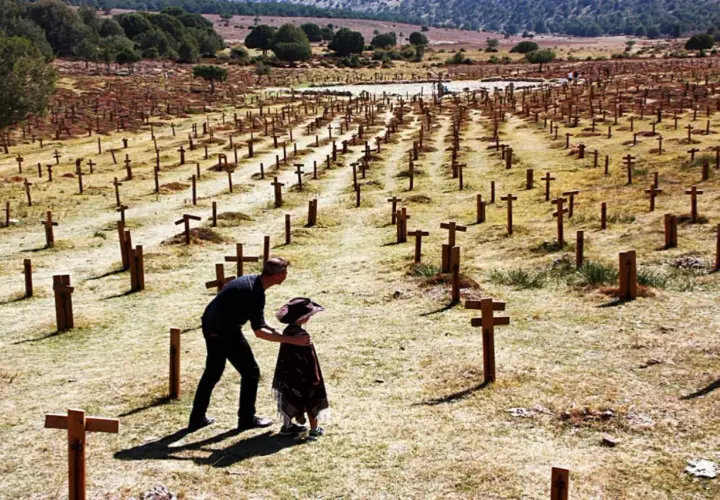 Un hombre y un niño observan un vasto cementerio sad hill, con hileras de cruces de madera bajo un cielo despejado.