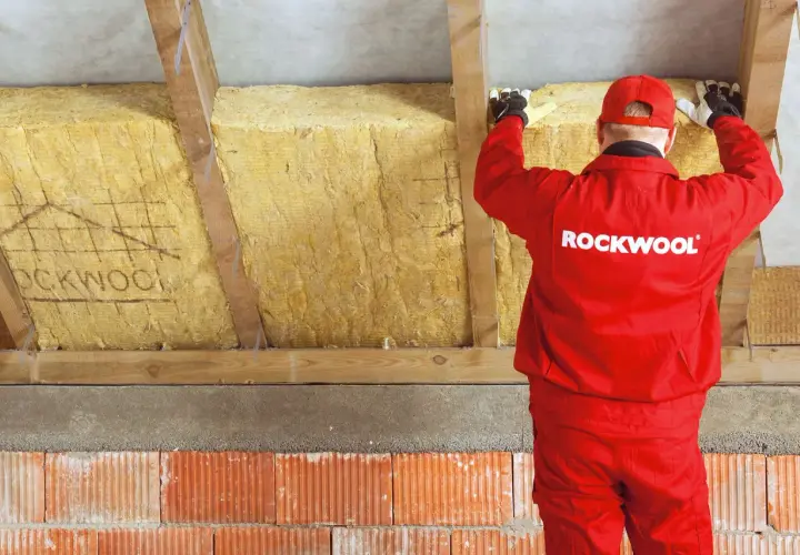 Pracownik w czerwonym stroju Rockwool montuje izolację z wełny mineralnej, tworząc zabudowę murłaty od zewnątrz.