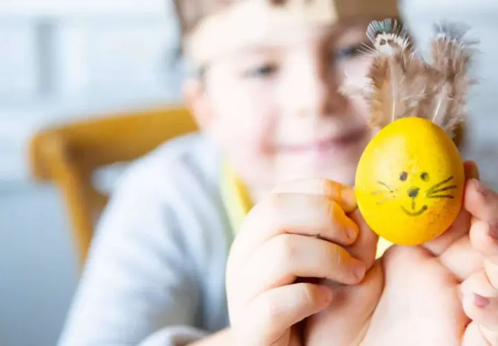 Ein Kind hält ein gelbes Ei mit lustigen Ostermotiv-Gesicht und Federn als Ohren.