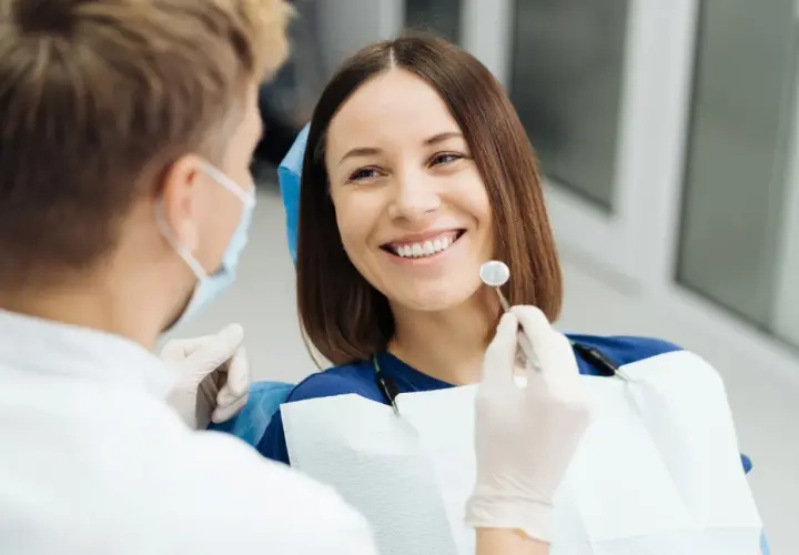 Uśmiechnięta pacjentka podczas wizyty u dentysty. Lekarz w masce i rękawiczkach bada jej zęby, przygotowując się do ewentualnego piłowania zębów.