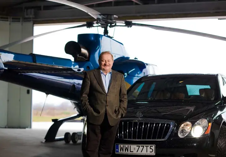Tadeusz Gołębiewski w garniturze, z rękami w kieszeniach, stoi przed helikopterem i luksusowym samochodem. Marzenia o podróżach i sukcesie, jak te, które mają dzieci, są tu namacalne.