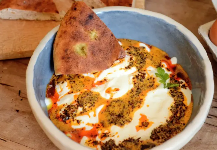 Un bol de cilbir recette, avec du pain pita trempé dans une sauce crémeuse et épicée, garni d'herbes fraîches.
