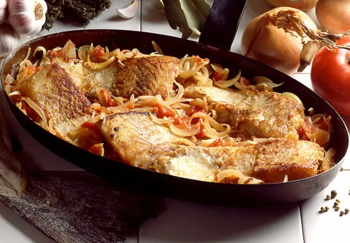 Morue à la lyonnaise, des filets de poisson dorés mijotant avec des oignons et des tomates dans une poêle.