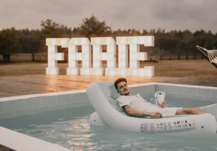 Mężczyzna relaksuje się na dmuchanym materacu w basenie, z wielkimi literami "FARTE" w tle. Idealne miejsce na kawalerski!