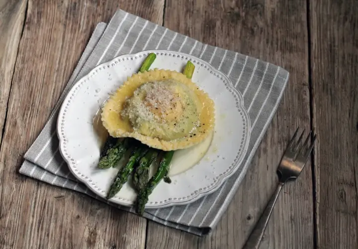 Ravioli z żółtkiem, podane z zielonymi szparagami i posypane parmezanem, na białym talerzu.
