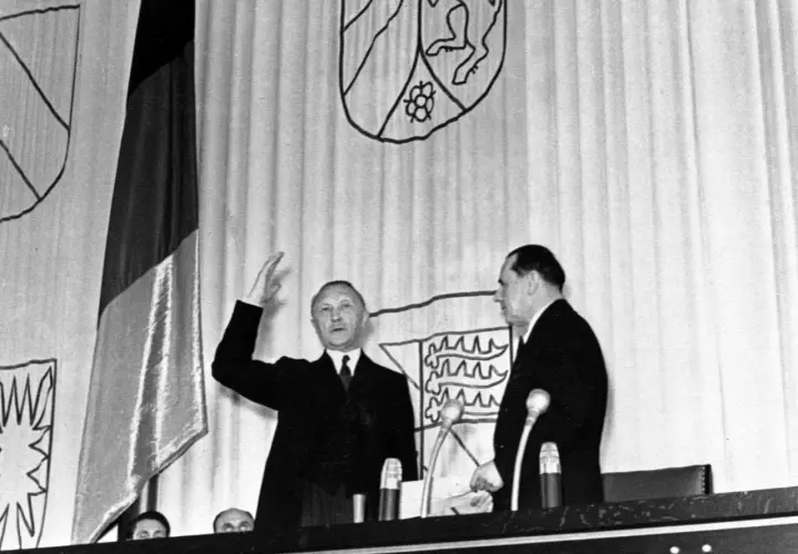 Ein Mann hebt die Hand, neben ihm ein weiterer Mann. Im Hintergrund die deutsche Fahne und Wappen. Der ältere Bundeskanzler spricht.