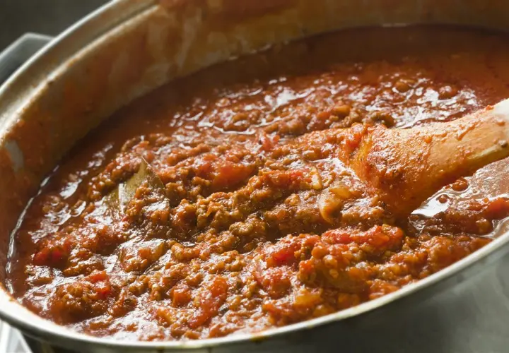 Salsa boloñesa burbujeante en una olla, con una cuchara de madera removiendo la carne picada y los tomates.