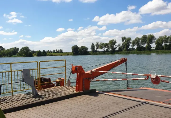 Prom na rzece, z widokiem na zielony brzeg i chmury. W oddali widać drzewa, a na promie pomarańczowy dźwig i znak STOP. To widok z okolic Piotrkowa Kujawskiego.