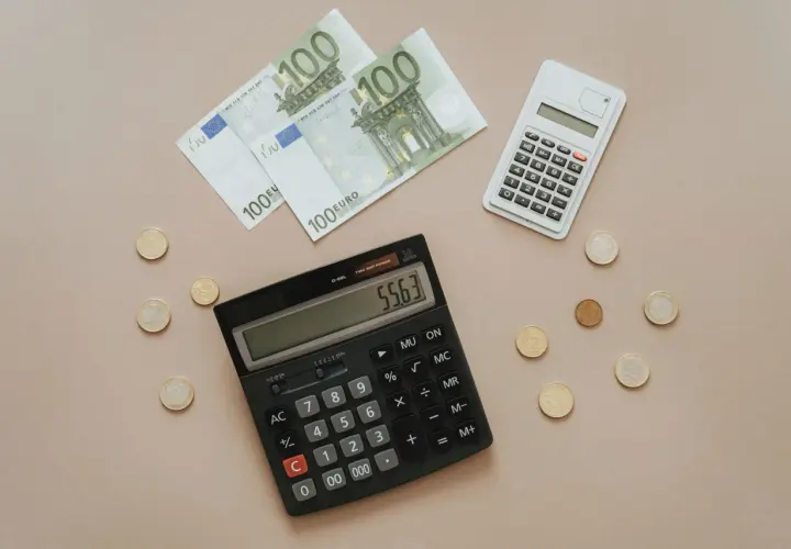 Calcolatrice e banconote da 100 euro, con monete sparse. Si sta calcolando quanto costa la tassa di soggiorno.