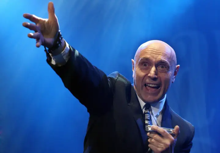 Artysta w garniturze, z podniesioną ręką, gra na harmonijce ustnej. Jego ekspresja sugeruje, że wykonuje energetyczne **shakin dudi utwory**.