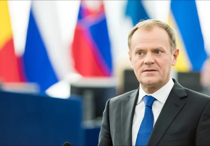 Donald Tusk w garniturze i niebieskim krawacie na tle flag.